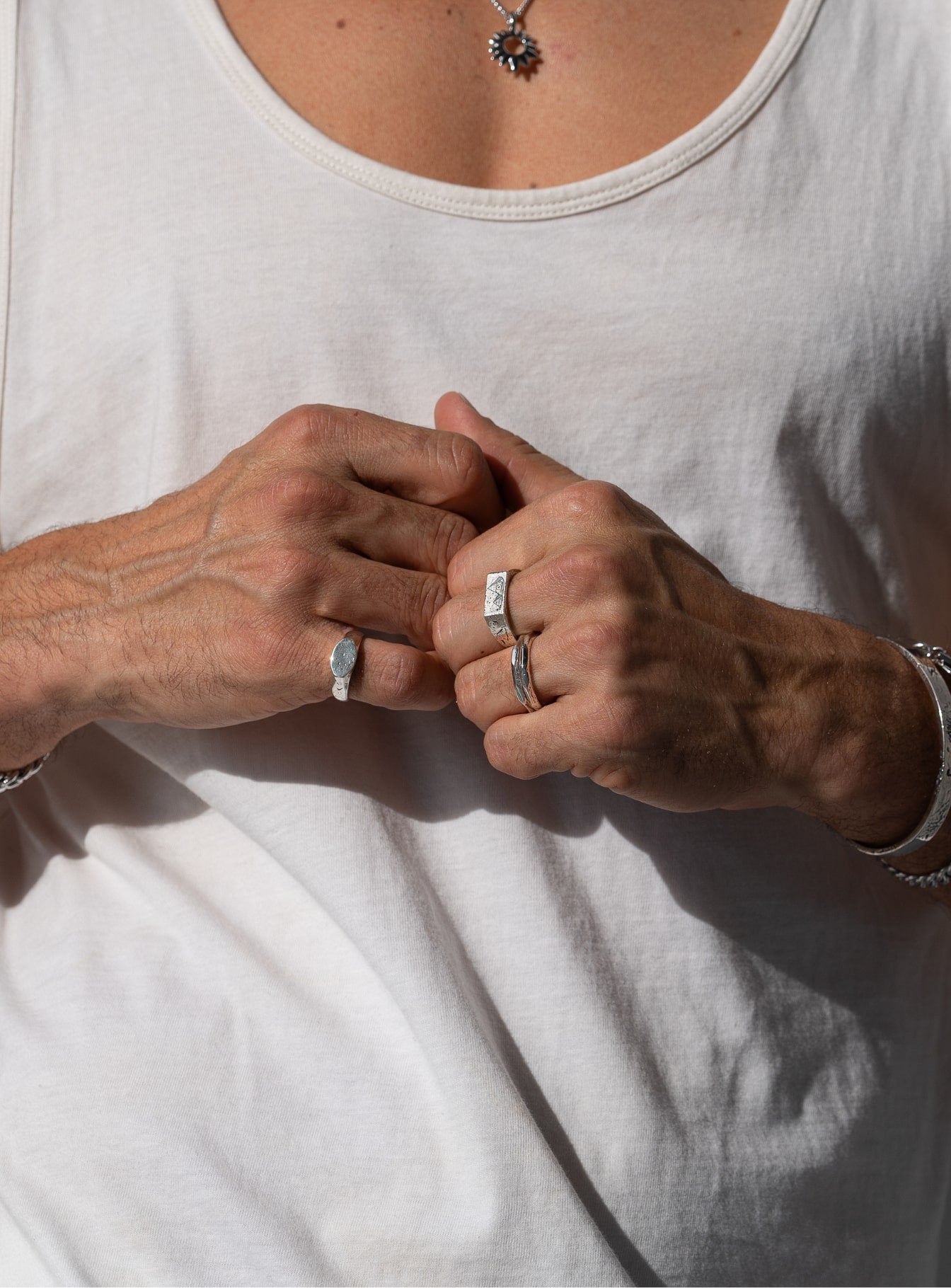 Man wearing silver rings and a sunburst pendant necklace, styled with a minimalist white tank top, showcasing a modern and rugged jewellery aesthetic.