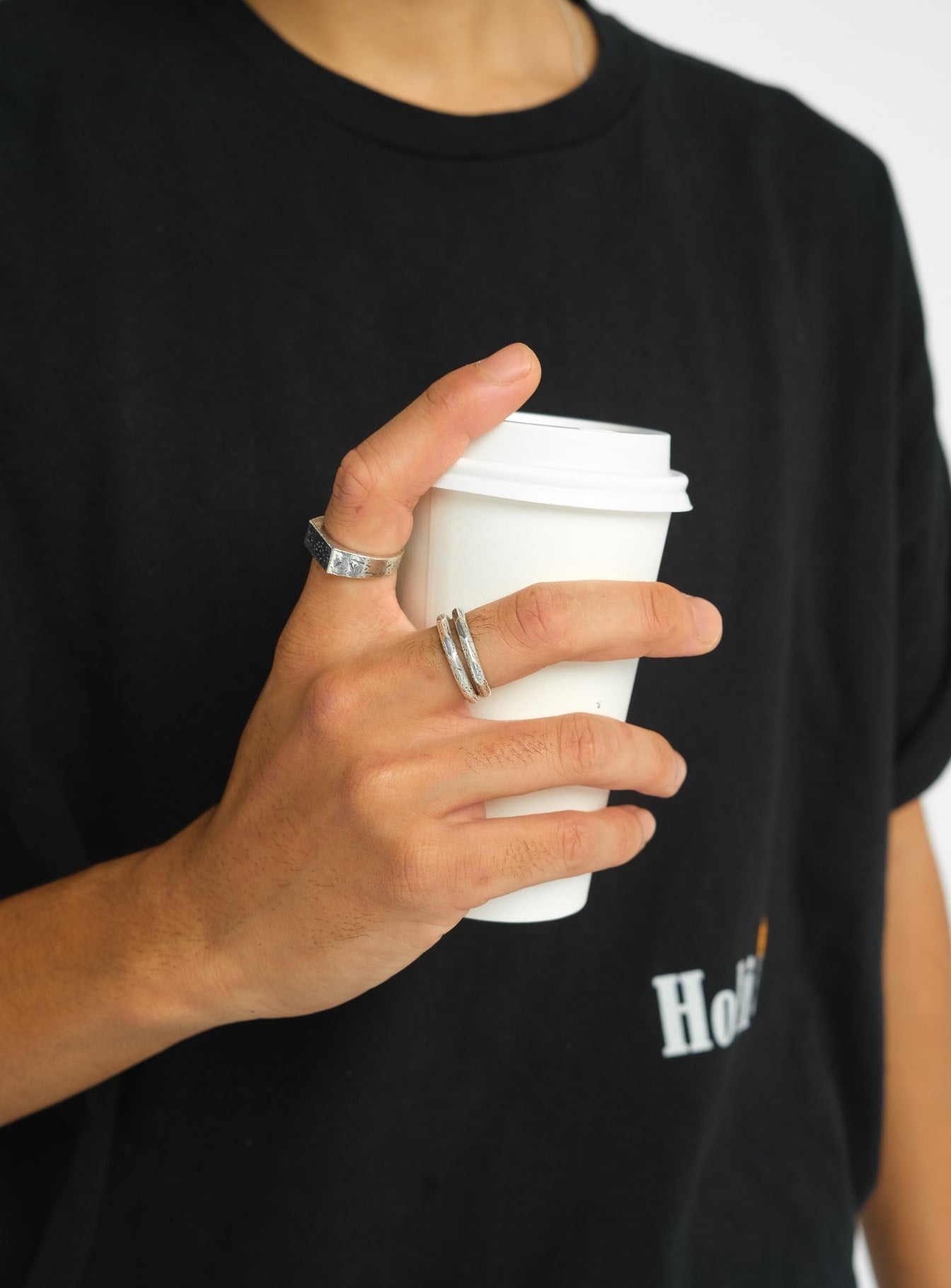 Close-up of a hand holding a white coffee cup, showcasing modern silver rings with a textured design, styled with a casual black t-shirt.